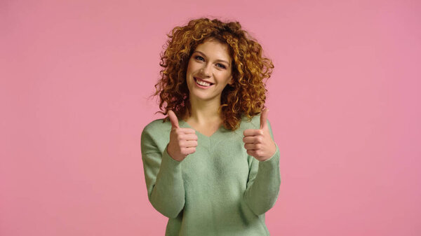 young and joyful woman with red hair showing thumbs up isolated on pink