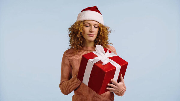 thoughtful woman in santa hat looking at large gift box isolated on blue