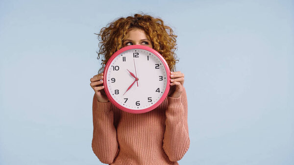 sly redhead woman looking aside while hiding face behind round clock isolated on blue