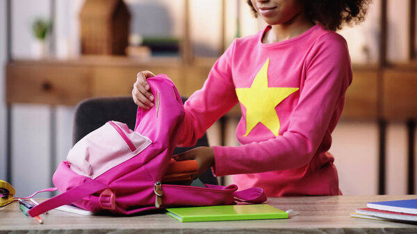 cropped view of african american child putting notebooks in backpack
