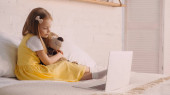 Preschooler child hugging soft toy near laptop on bed at home 