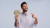 happy man in shirt looking at camera while pointing at dollars isolated on blue 