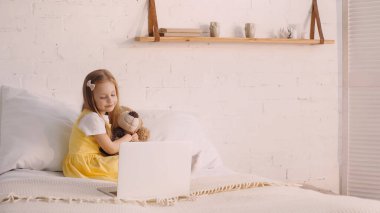 Smiling kid hugging teddy bear near laptop on bed  clipart