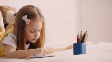 Anaokulu çocuğu evdeki yatakta kağıda resim çiziyor. 