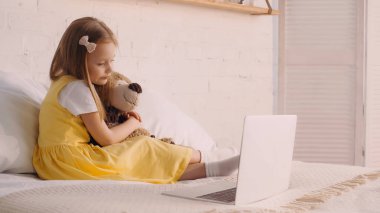 Anaokulu çocuğu evde laptopun yanında yumuşak oyuncağa sarılıyor. 