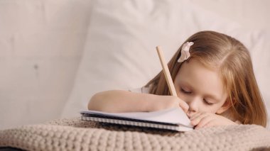 Çocuk yatak odasında battaniyeye uzanmış resim çiziyor. 