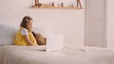 Kid looking at laptop near soft toy on bed at home  clipart