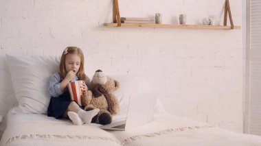 Kid eating tasty popcorn near soft toy and laptop in bedroom  clipart