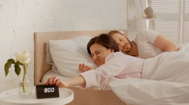 Displeased woman turning off clock near sleeping husband at home  clipart