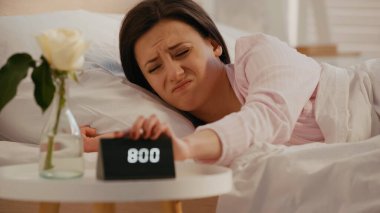 Displeased woman turning off clock near blurred plant in bedroom  clipart
