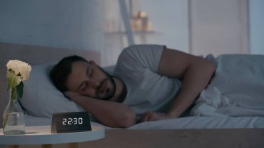 Evde uyuyan bulanık adamın yanındaki vazoda saat ve çiçek. 