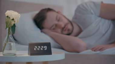 Evde uyuyan adamın yanında, komodinin üzerinde gül ve saat. 