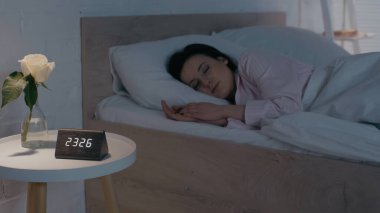 Evde uyuyan bir kadının yanındaki komodinin üzerinde çiçek ve saat. 