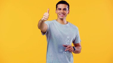 joyful young man in t-shirt holding bottle with water and showing thumb up isolated on yellow  clipart