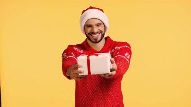 Noel Baba şapkalı gülümseyen adam sarı kutuda izole edilmiş bir hediye kutusu tutuyor.