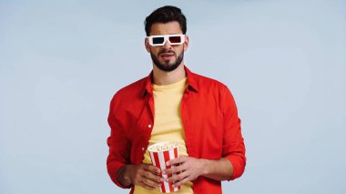 Üç boyutlu gözlüklü hoşnutsuz ve sakallı bir adam film izliyor ve elinde patlamış mısır kovası var. 