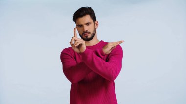 Pembe kazaklı ciddi bir adam ellerini çapraz yapıp mavi üzerine parmak işareti yapıyor. 
