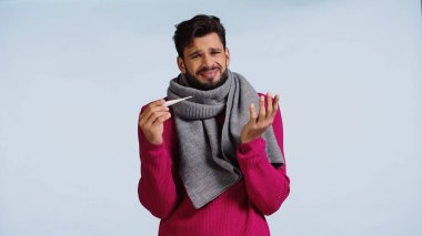 Pembe kazaklı ve atkılı hasta adam mavi üzerinde dijital termometre tutuyor. 