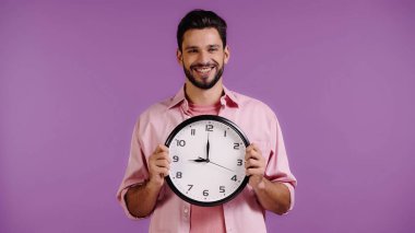 Pembe gömlekli mutlu genç adam elinde mor bir saat tutuyor. 
