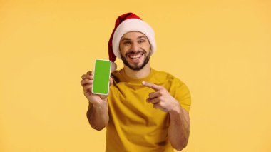 Noel Baba şapkalı neşeli adam sarı ekranda yeşil ekranlı akıllı telefonu işaret ediyor.