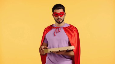 Süper kahraman kostümlü sakallı adam sarı kutuda izole edilmiş pizza kutusu tutuyor.