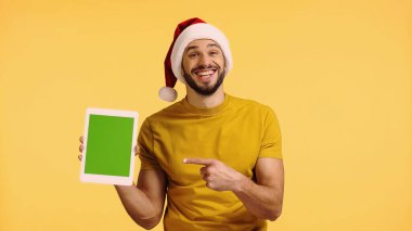 Noel Baba şapkalı neşeli adam sarı tablet üzerinde izole edilmiş dijital tableti işaret ediyor