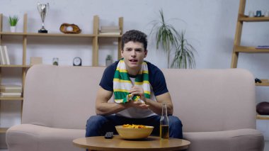 excited sport fan sitting on sofa near chips and beer and watching game on tv clipart
