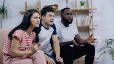 Memlekette spor müsabakası izleyen gergin arkadaşlarının yanında elleri işaret eden hoşnutsuz Afro-Amerikan bir adam.