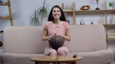 Mutlu spor fanatiği kadın televizyonda basketbol maçı izlerken kanepede topuyla oturuyor.