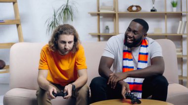 KYIV, UKRAINE - 22 Aralık 2021: Üzgün arkadaşına bakan gülen Afro-Amerikan adam joystick ile kanepede oturuyor.
