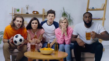 Afro-Amerikan bir adam elinde bir kupa bira tutarken evde arkadaşlarıyla futbol maçı izliyor.