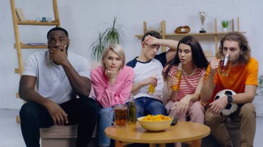 Genç adam futbol topuyla bira içerken aynı zamanda çok ırklı arkadaşlarla futbol maçı izliyor.