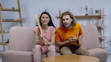 Mutlu kadın elinde beysbol sopası ile TV 'de maç izlerken şaşkın arkadaşının yanında.