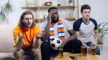 Evde ırklar arası arkadaşlarla futbol maçı izlerken elleri gösteren üzgün bir adam.