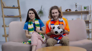 Futbol topuyla neşeli bir adam televizyonda endişeli bir kadının yanında şampiyonluk maçı izliyor.