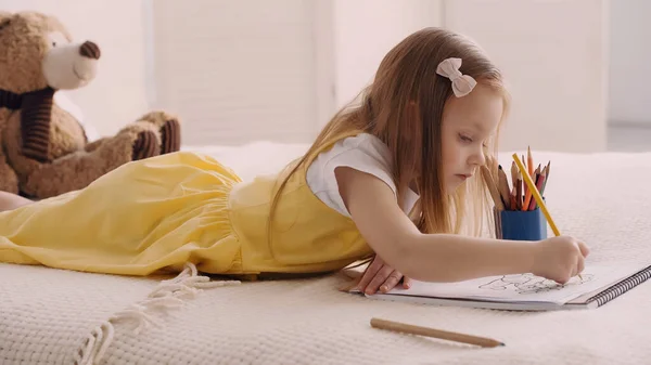 Child drawing with color pencil near teddy bear on bed