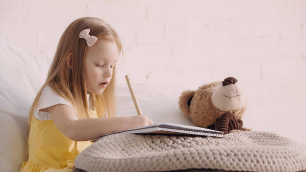 Preschooler kid drawing on paper near teddy bear and blanket at home 