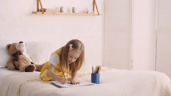 Child drawing on paper near color pencils and soft toy on bed