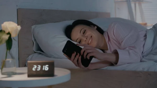 Mujer Alegre Usando Teléfono Inteligente Cerca Reloj Borroso Planta Dormitorio — Foto de Stock