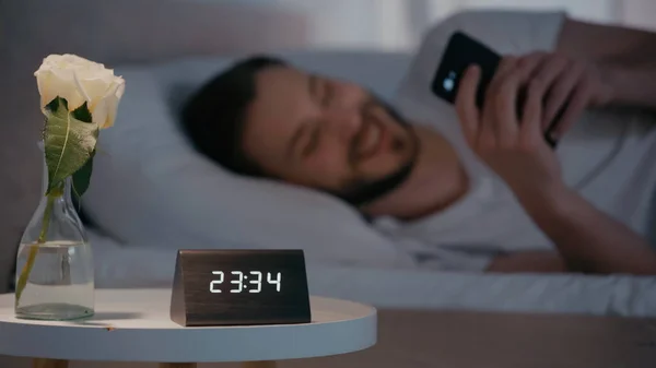 夜にベッドの上でスマートフォンを使用して 男の近くのベッドサイドテーブルの上に時計とバラ — ストック写真