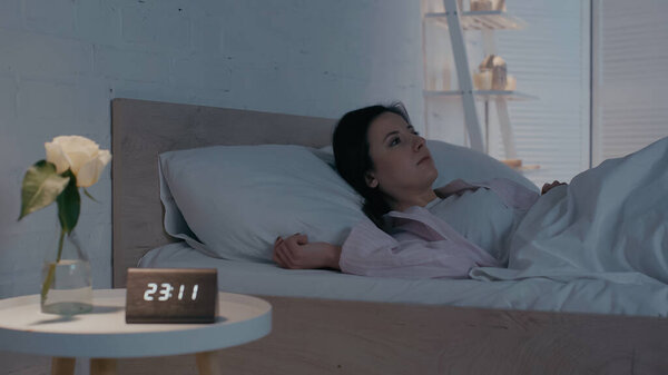 Woman lying on bed near blurred clock and plant on bedside table at night 