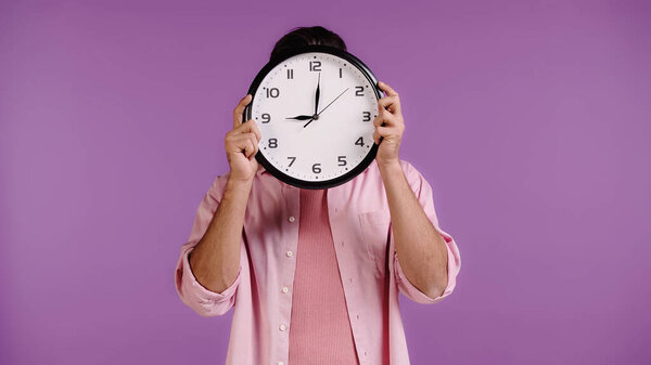 young man obscuring face with clock isolated on purple