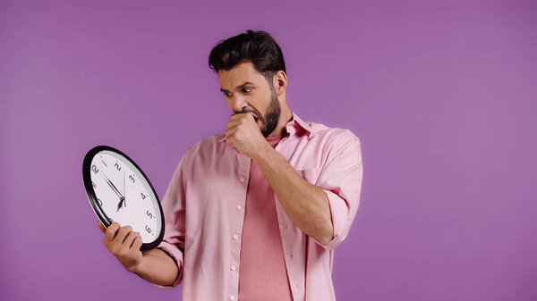 worried young man looking at clock isolated on purple 