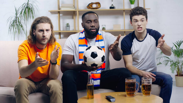 frustrated multiethnic football fans pointing with hands while watching championship on tv