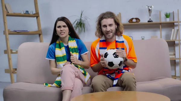 cheerful man with soccer ball watching championship on tv near worried woman