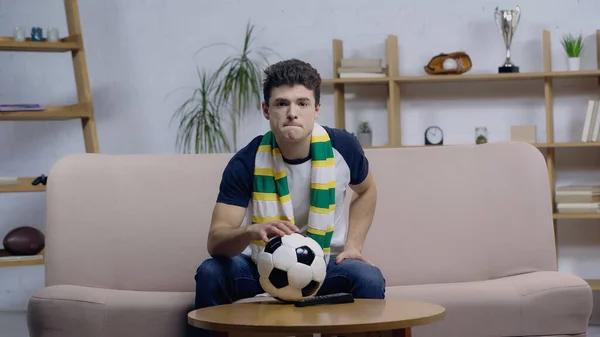 nervous sport fan in striped scarf watching football match near ball on coffee table