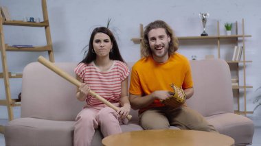 Kızgın kadın elinde beyzbol sopasıyla mutlu bir adamın yanında TV izlerken.