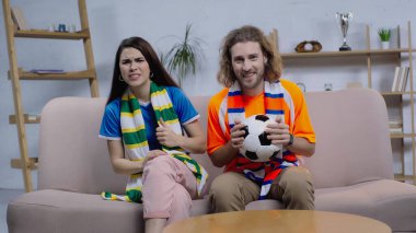 TV 'de gergin bir kadının yanında futbol topu tutan gülümseyen bir adam.