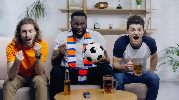 african american man screaming and showing win gesture while watching football match with excited friends