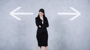 pensive businesswoman in suit standing near arrows on grey 